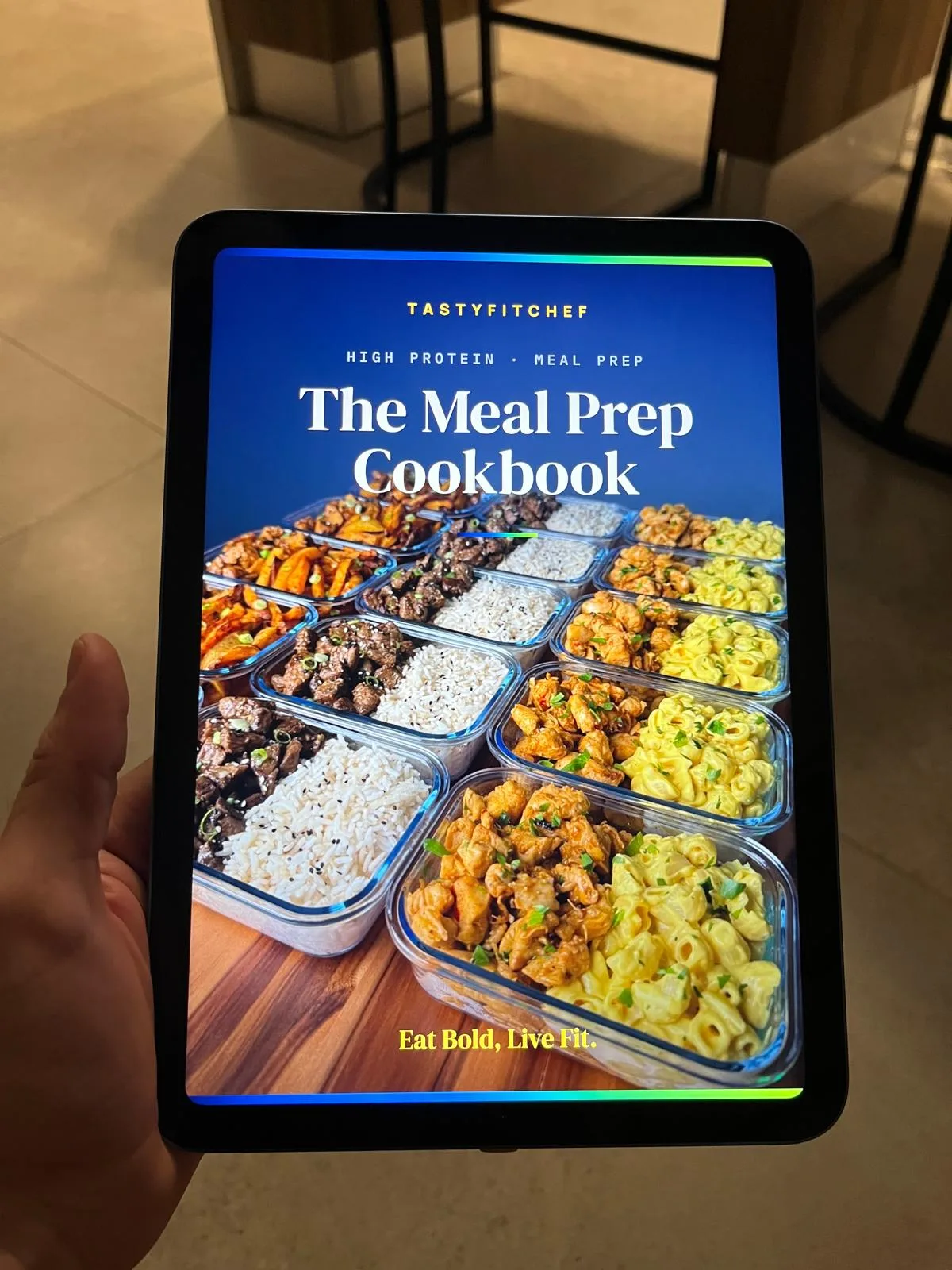 Hands holding an iPad showing the cookbook, kitchen in the background