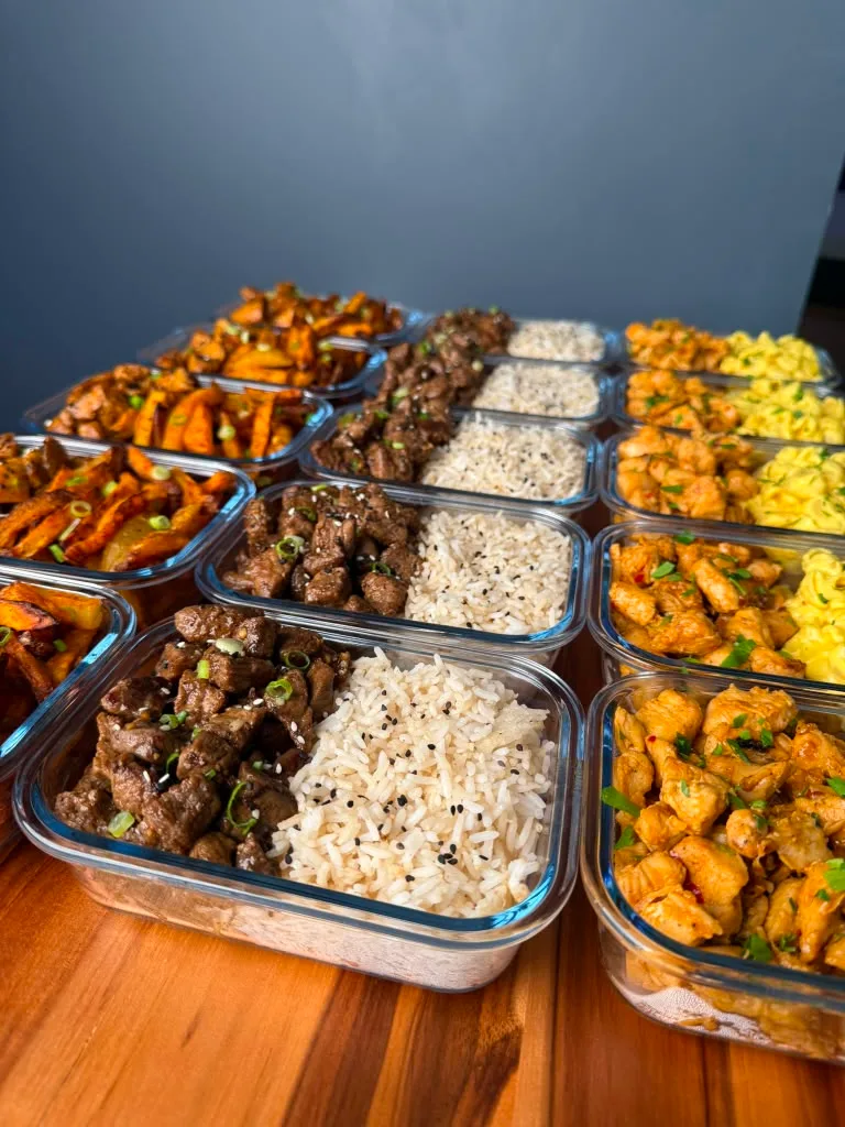 Fifteen glass containers of prepped high-protein meals on an acacia board, from above