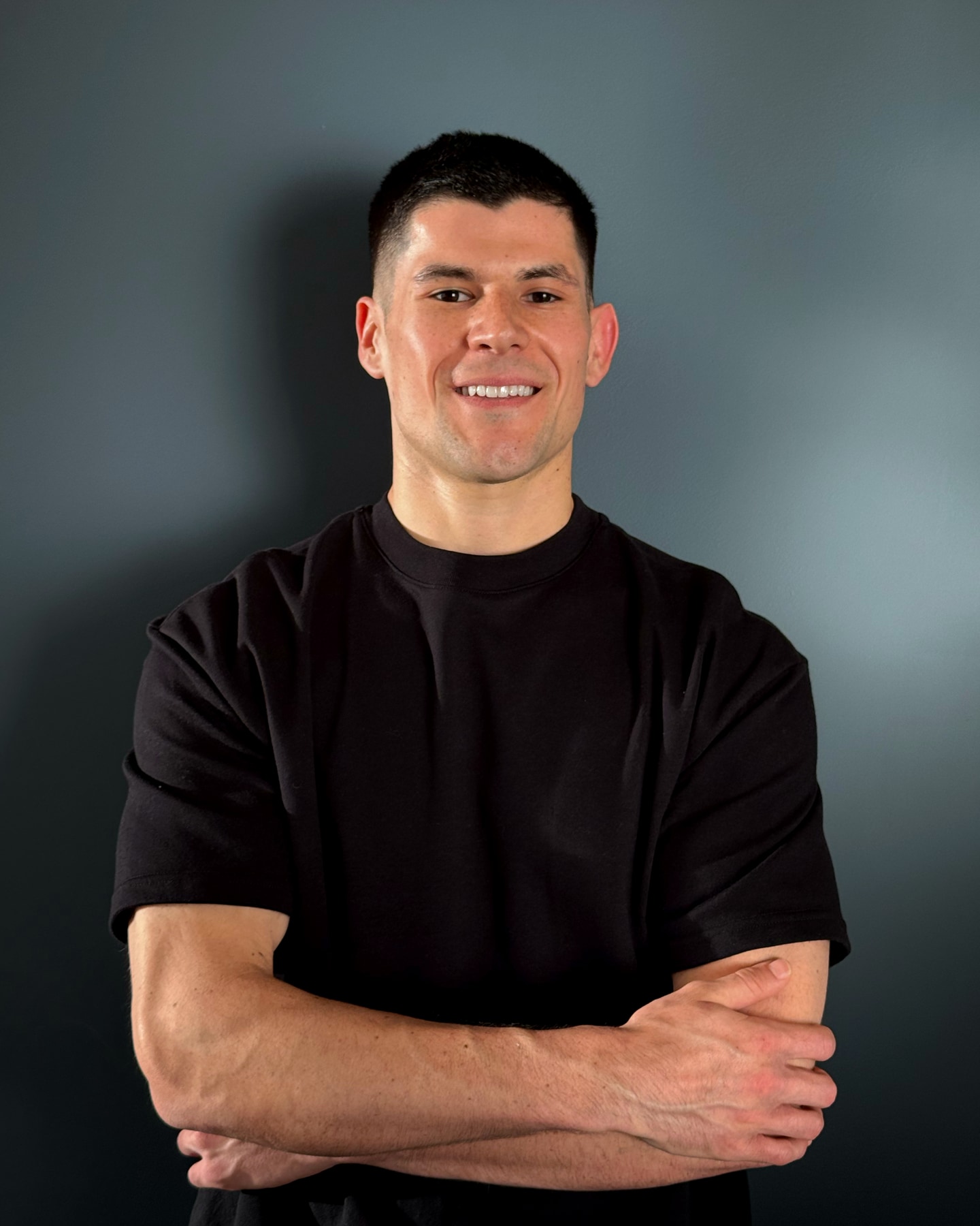 Ruben, founder of TastyFitChef, smiling with his arms crossed in a black t-shirt against a grey studio backdrop